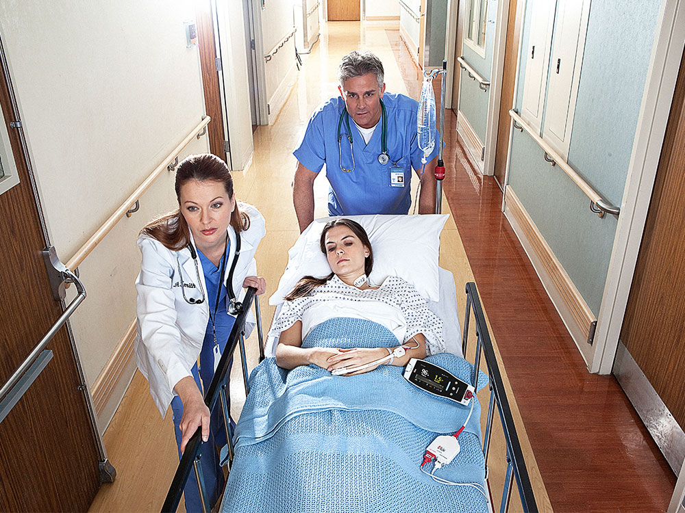 Dark-skinned patient in hospital bed monitored by Masimo Root device.
